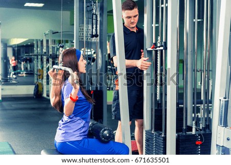 Beautiful sportive woman in sportswear doing exercise with a heavy weight with her trainer using training apparatus in a gym
