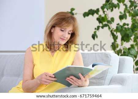 Attractive mature woman sitting on sofa at home and reading magazine