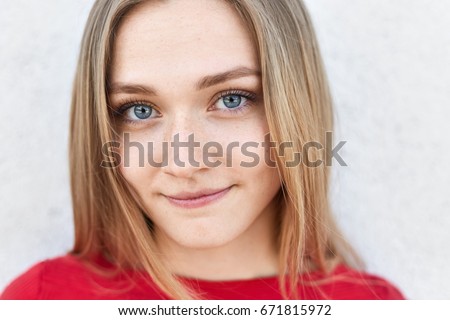 Close up portrait of pretty female with blue alluring eyes, freckled face and pure healthy skin looking directly into camera having thoughtful expression. Cute young female in red clothes posing Royalty-Free Stock Photo #671815972