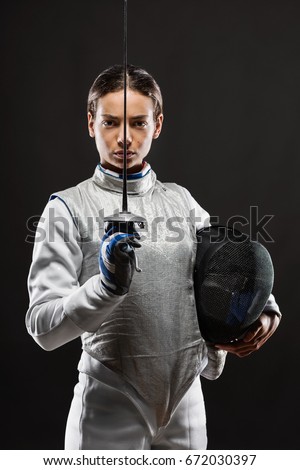 Portrait of Young woman fencer wearing white fencing costume and holding sword and mask. Looking at camera. Black Background Royalty-Free Stock Photo #672030397