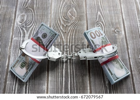 Bills of one hundred dollars in handcuffs, like hands. In the background there is a table made of wood. Idea: Detention of an attacker, trial, release on bail, arrest, theft of money, bribe. Royalty-Free Stock Photo #672079567