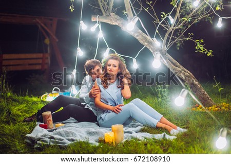 Young couple in magical night forest decorated light garlands. Sitting on plaid and kiss each other. 
Man and woman hugs. 