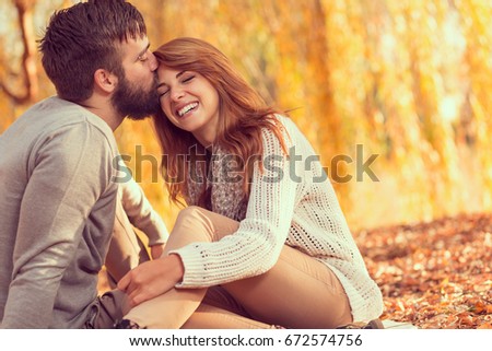 Couple in love sitting on autumn fallen leaves in a park, enjoying a beautiful autumn day. Man kissing a woman in a forehead 