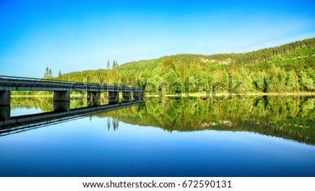 Summer landscape with trees and bridge reflected in the lake water. The picture taken at the lake Selbu, Norway.