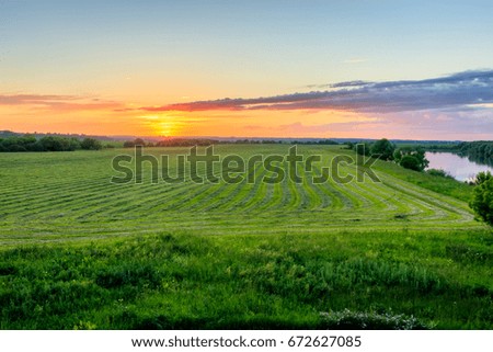 Twilight at cultivated land in the countryside on a summer evening with cloudy sky background and river. Procurement of food for animals. Combined field. Landscape.