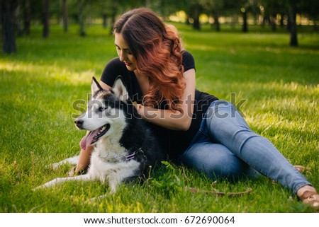 A beautiful girl is playing with her dog. Siberian husky dog with blue eyes. Bright green trees and grass are in the background. Friendship between man and animals