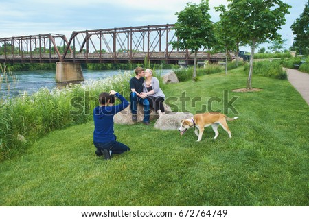 Woman taking photo of happy young Caucasian couple sitting together on stones with bridge behind them. Man kissing his girlfriend, their dog walking nearby