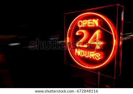 Open 24 Hours neon sign on the road side at night with moving cars.