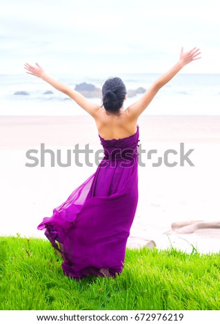 Beautiful biracial Asian Caucasian teen girl wearing purple magenta dress standing on grassy knoll overlooking ocean, arms raised