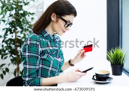 Beautiful girl with black hair wearing shirt and eyeglasses sitting in cafe with mobile phone and cup of coffee, holding credit card, online shopping.