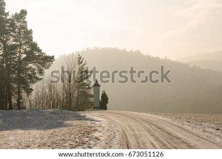 Scenic panoramic foggy color winter photography of road with tracks and a memorial cross in a winter idyllic hilly landscape with snow on,  sunny diffuse light,  twilight