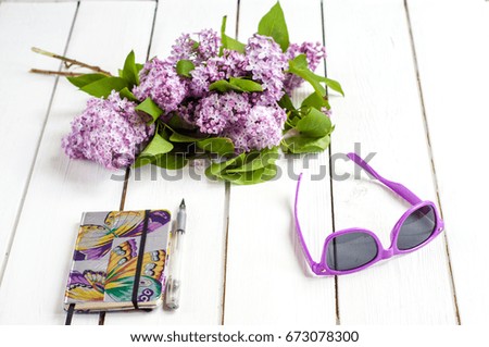 Lilac bouquet on white wooden background, silver pen and purple notebook, purple sunglasses