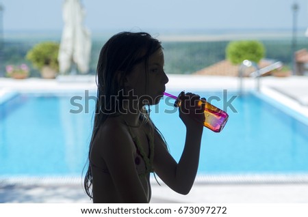 Little child girl sitting in front of pool and drinking on the straw from bottle . Sitting on table and enjoy. Refreshment  in summer day on school holiday vacation.
