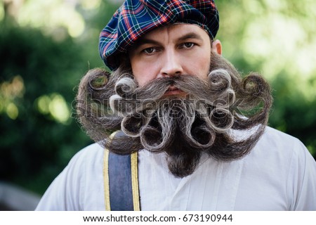 Close-up portrait of a brave Scot with a amazing beard and mustache curls . Checkered red Scottish kilt skirt, hat with pompon, cane and sword. Beard styling by professional barbershop. Royalty-Free Stock Photo #673190944