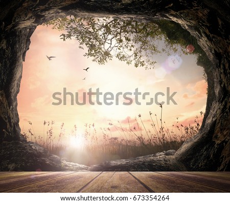 He is risen concept: Wooden table with open empty tomb stone on meadow autumn sunrise background Royalty-Free Stock Photo #673354264