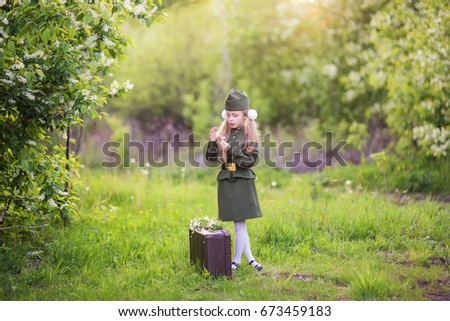 Victory Day. A girl in soldier's clothes stands next to a suitcase in the grass under a tree