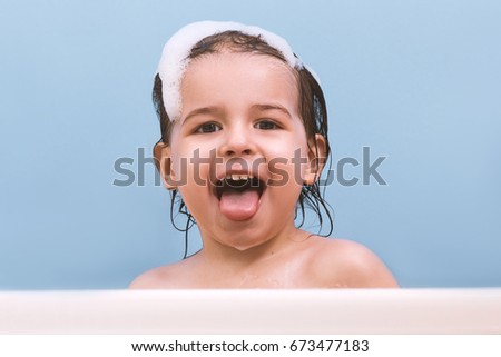Fun cheerful happy toddler baby taking a bath playing with foam bubbles. Little child in a bathtub showing tongue. Smiling kid in bathroom on blue background. Toddler washing and bathing. Health care.
