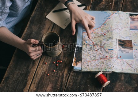 Woman's hand marks route on paper map of Europe using pins and red rope. Another hand holds cup of tea. Girl gets inspired with photos of nature and planning journey on dark wooden table Royalty-Free Stock Photo #673487857