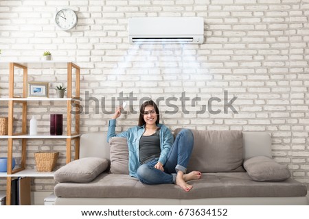 Happy Young Woman Holding Remote Control Relaxing Under The Air Conditioner Royalty-Free Stock Photo #673634152