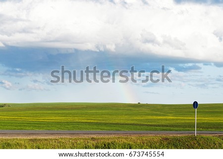 Green meadow under blue cloudy sky with a rainbow after rain afar. Rural road and road sign in the foreground.