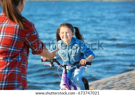 Young woman teaching her daughter to ride bicycle outdoors near river