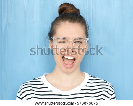 Close up portrait of a happy young hipster european teen girl in nerdy square glasses blinking at camera in a playful manner, smiling and winking, shouting loudly, standing against blue background