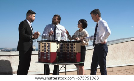 Group of engineers and architects in a meeting standing outdoors on a pation discussing a model of office or apartment blocks in a low angle view against blue sky.