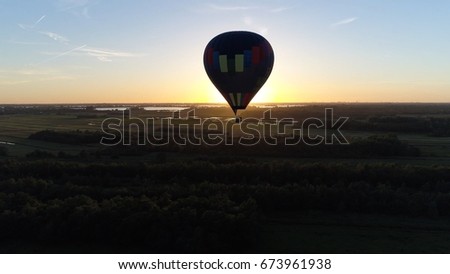 Aerial bird view photo near hot air balloon in flight during sundown aircraft over nature reserve meadows and pastures in far background warm colors of sundown sunlight showing on horizon