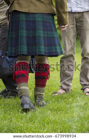 The feet of a man in the military uniform of a Scottish warrior. In kilt, leggings and boots against the background of green grass, close-up