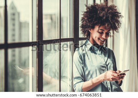 Beautiful Afro American girl in casual clothes is using a smart phone and smiling while standing near the window Royalty-Free Stock Photo #674063851