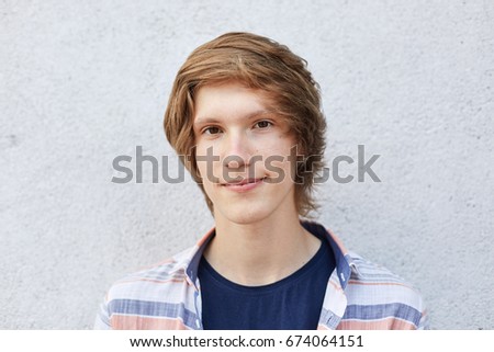 Close up of handsome male with dark eyes, pure healthy skin and thin lips looking directly into camera with serious expression. Hipster boy with appealing appearance posing against white wall