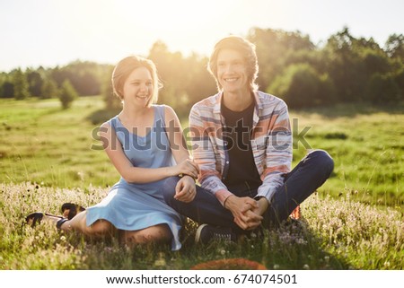 Outdoor portrait of happy girl with bobbed hair dressed in blue dress sitting near her friend having fun togehter enjoying beautiful nature. Stylish teenage boy sitting crossed legs near his friend