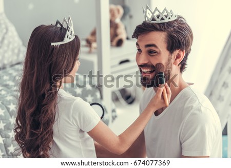 Cute little daughter and her handsome young dad in crowns are playing together in child's room. Girl is doing her dad a makeup