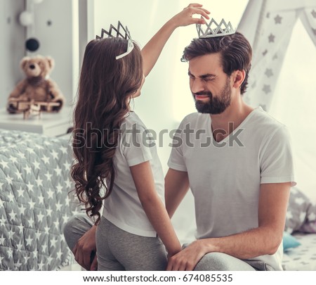 Cute little girl is putting a crown on her handsome young dad's head and smiling while they are playing together in child's room