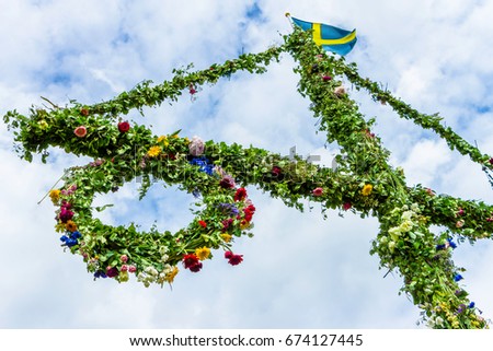 A pole and flag against blue sky and white clouds. A maypole decorated, covered in flowers and leaves.  Royalty-Free Stock Photo #674127445