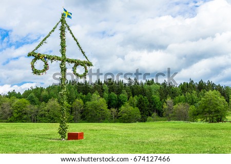 A pole and flag against green trees and blue sky. A maypole decorated, covered in flowers and leaves.  Royalty-Free Stock Photo #674127466