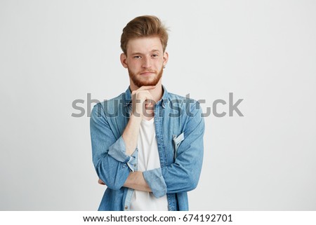 Portrait of confident young handsome man with beard looking at camera thinking with hand on chin over white background.