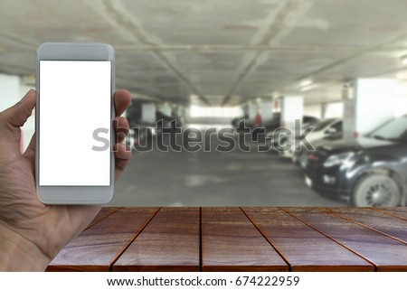 Empty wooden table space platform and Hand holding smartphone with white blank screen over blurred  Parking lot in the mall background for product display montage.
