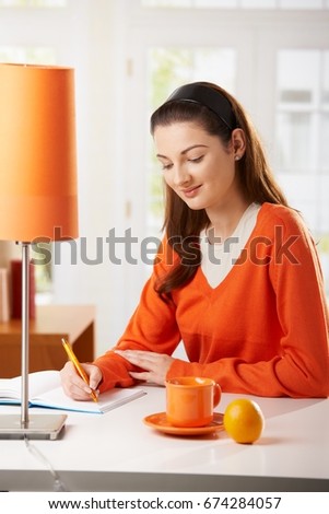 Young woman sitting at desk at home, writing notes to diary, looking down, smiling.