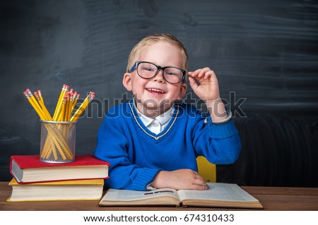 Happy cute clever boy is sitting at a desk in a glasses with raising hand. Child is ready to answer with a blackboard on a background. Ready for school. Back to school. Apple and books on desk Royalty-Free Stock Photo #674310433