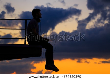 Silhouette of a man against background of clouds and golden sunset. He sits on edge of roof. The guy is holding a tablet PC.