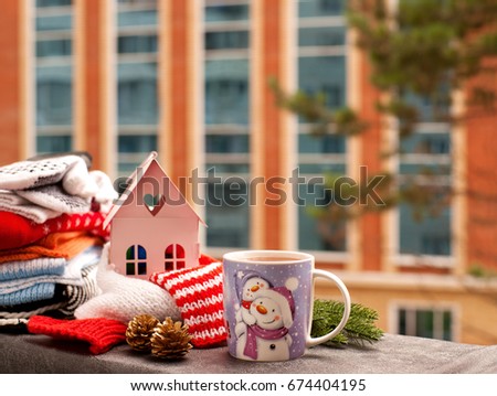 Tea and New Year. A mug of tea with a picture of a snowman on the balcony. A knitted scarf shrouds the house and creates a cosiness for the new year. Warm winter clothes on the window sill.