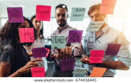 Business investors discussing work looking at the information on sticky notes stuck in office. Colleagues standing in front of post-it notes stuck on glass and discussing.