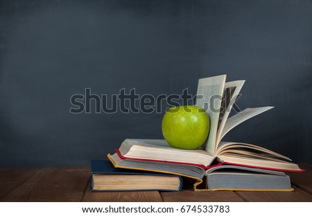Back to school background. Green apple lies on the pile of open books on the wooden desk. Blackboard as a background. 