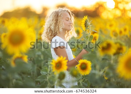 Pretty curly girl in white dress in field of sunflowers over sunset lights