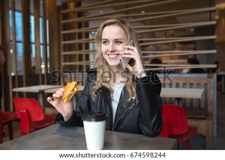Sweet blonde in a leather jacket at a fast food cafe eating a hamburger, drinks coffee and talks on the smartphone with a smile