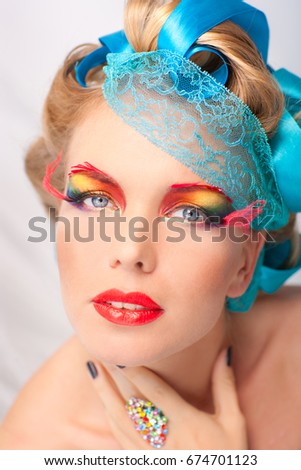 Studio photograph of a beautiful young woman with blond hair decorated with blue ribbons. With artistic colorful makeup. Photo is retouched. 