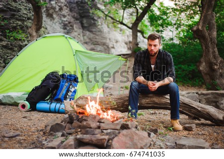 Traveler man relaxing, looking at the fire and dreaming at camping tent outdoors on nature. 