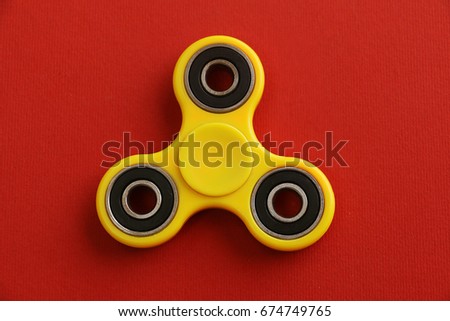 Yellow Fidget Spinner on red background  is useless machine designed to spin but became a popular toy for people who wanted to cut boredom and stress. Banned in schools due to distracting nature.