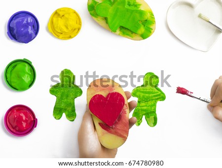 Childs hand, play painting with potato stamps, brushing green on a figure shape, red ,yellow, and purple poster paints  some isolated objects on a white background, copy space 